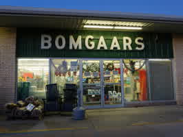 A photo of the front of a Bomgaars hardware store. The storefront consists of beige stone and green sheet metal panelling, with glass doors in the middle and a lit uppercase plastic sign above the doors.