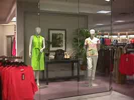 A display case in Dillards. A manequin is wearing a neon green summer dress, while another manequin is wearing white pants with a baby pink tropical shirt.