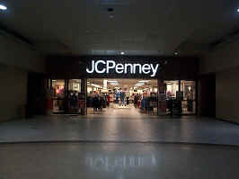 The front of the JCPenney. Walls are made of a brown fabric, with windows to the sides of the store entrance. The store sign is reflected in the mall's brown tile flooring.