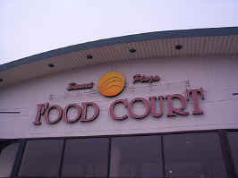 A close-up of the Sunset Plaza Mall sign. "Sunset Plaza" is in cursive, with large serif "FOOD COURT" letters below. A yellow circle representing the sun is in the middle, behind some wavy neon.