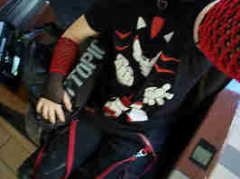 Me sitting in a mall massage chair. I'm wearing a Shadow the Hedgehog t-shirt, black and red bondage pants, black and red fishnet armwarmers, and a heart choker. I'm holding a Hot Topic bag in my right arm, with my left arm holding the camera.