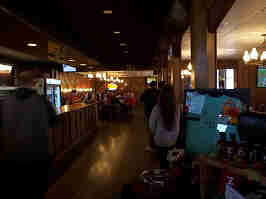 The inside of Zarda BBQ. Soft white lamps hang from a tiled ceiling, with wood floors, an order counter, and tables below.