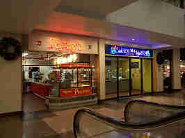 The storefronts of Topsy's and Curry In A Hurry. Both storefronts appear to be from the 80's, as both have neon signs, brown tile floors, and are lit by soft white and fluorescent lights.