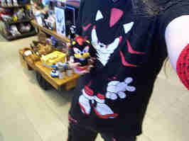Me, dressed in a Shadow the Hedgehog t-shirt, black and red bondage pants, and black and red armwarmers. I'm holding a small Shadow the Hedgehog plush with my right hand, and taking a photo with my left. I name this photo "me and the friend i got by being autistic".