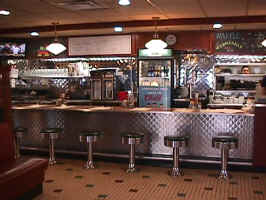 The bar at the Philips Avenue Diner. The bar has a brown-textured top with silver siding, the chairs are bar stools with brown vinyl cushioning and silver accents. The kitchen window and a fridge for drinks is in the background