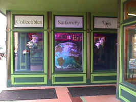 The side wall of an enterance to an antique store. Windows line the wall, giving a glimpse to the black-light lit insides. Inside is a globe and two hanging objects. The outside is painted dark green with light green accents.
