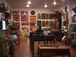 A room in an antiques store, housing wall-mounted collectibles. Old pinball tables sit in the midle of the room.