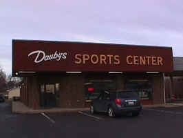 The front of Dauby's Sports Center. The storefront is made of brick, with a large sign consisting of a woodgrain background, white cursive "Daubys" text,  and light brown sans-serif "Sports Center" text.